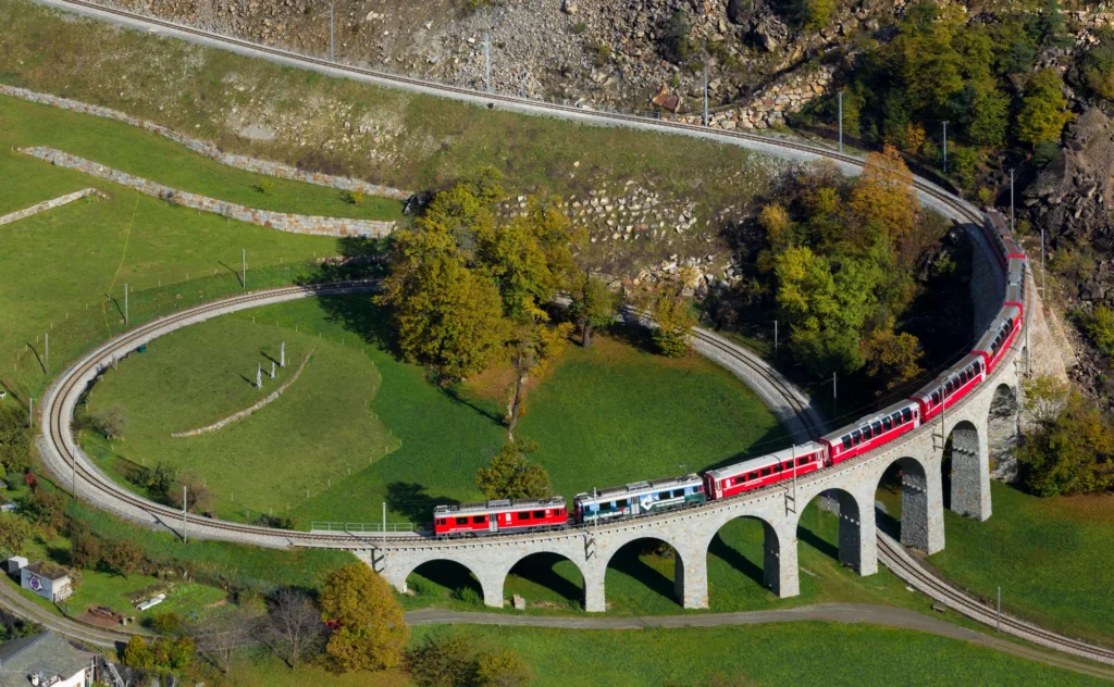 Red Rhaetian Railway train curving over the stone Brusio spiral viaduct between green meadows