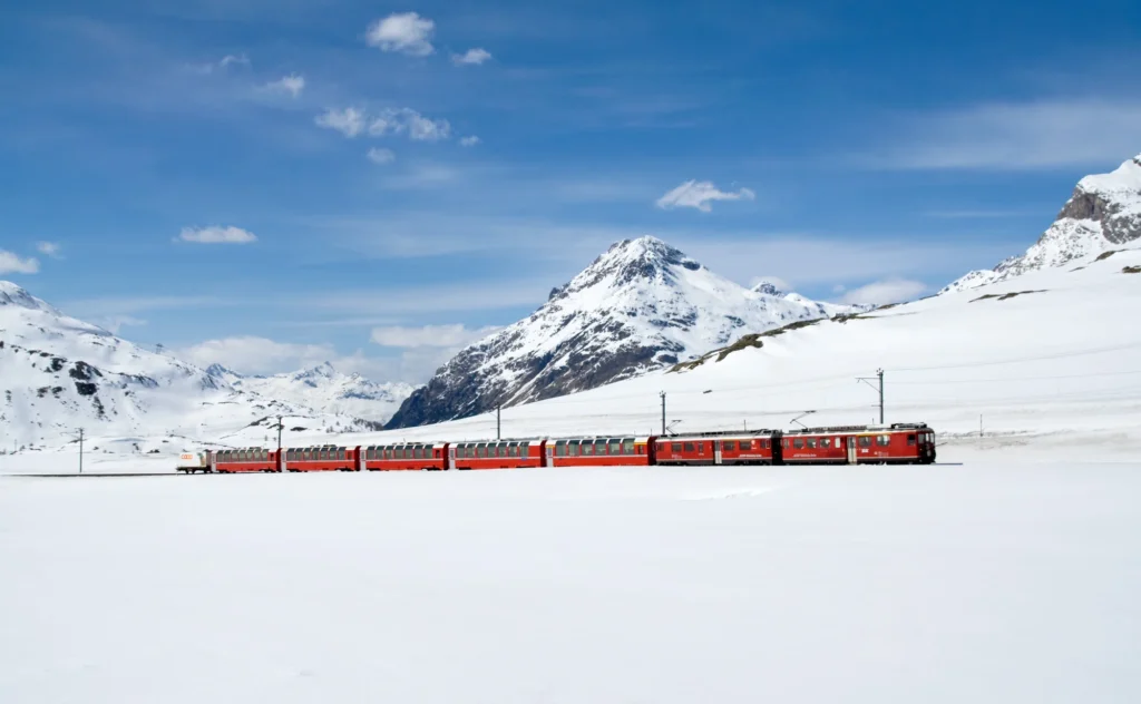Roter Bernina Express auf einer völlig verschneiten Hochebene am zugefrorenen Lej Pitschen, dahinter Berge