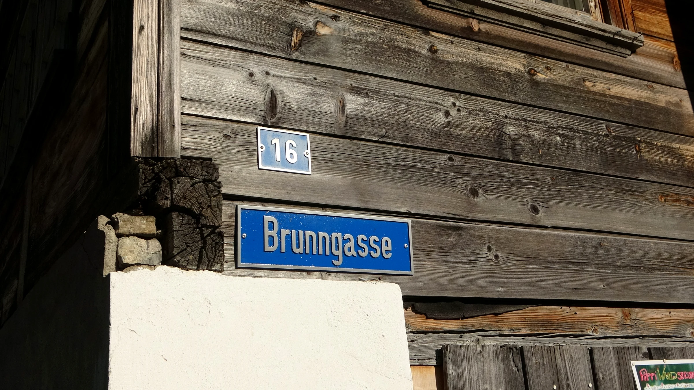 Blaues Straßenschild Brunngasse und Hausnummer 16 an einer alten, verwitterten Holzwand