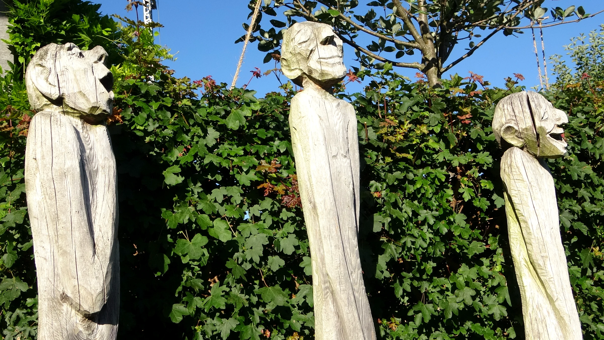 Drei hohe geschnitzte Holzfiguren mit Gesichtern vor einer Hecke in Brienz
