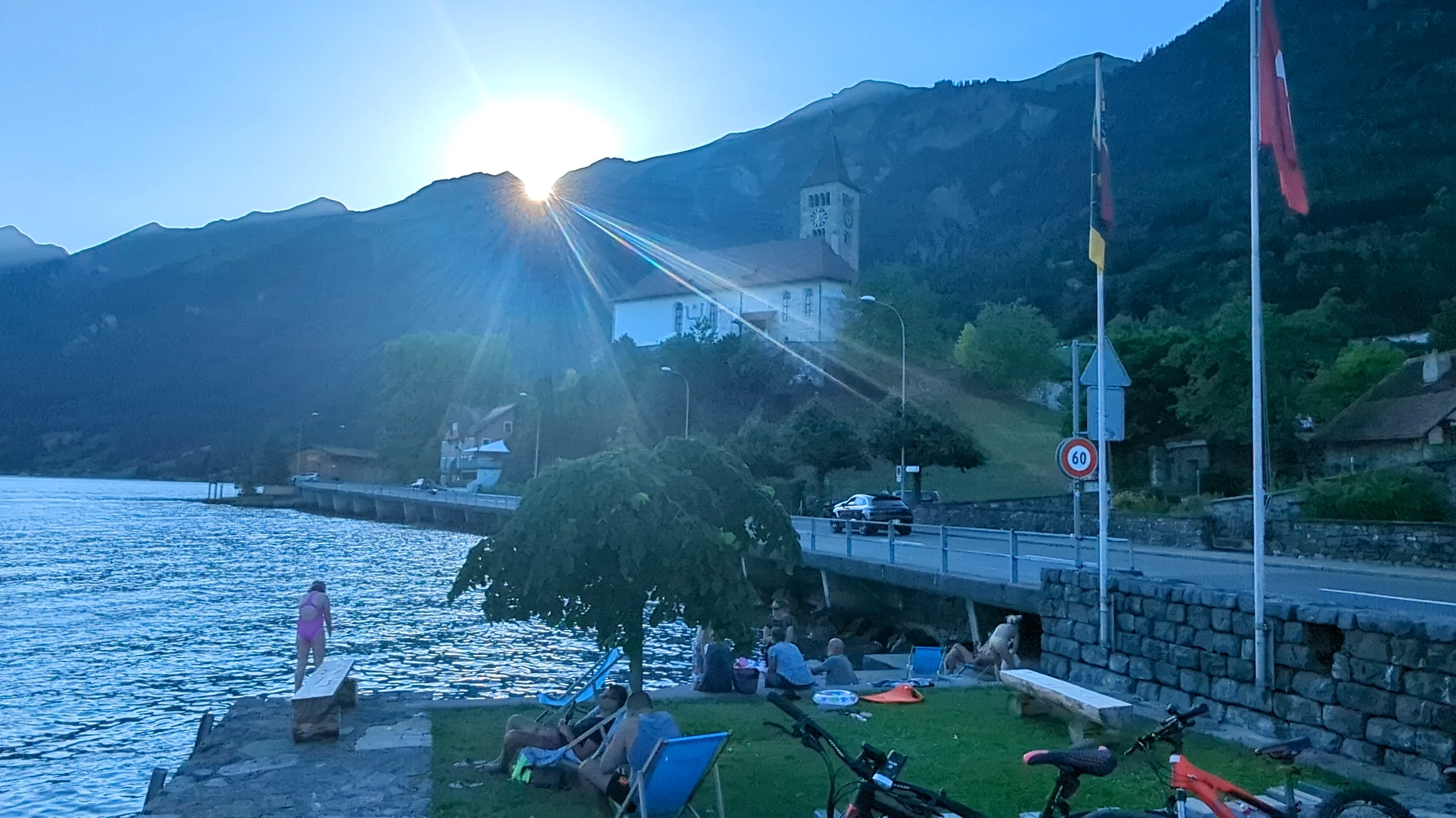 Abendsonne verschwindet hinter den Bergen über der Kirche von Brienz, Leute auf der Wiese am See