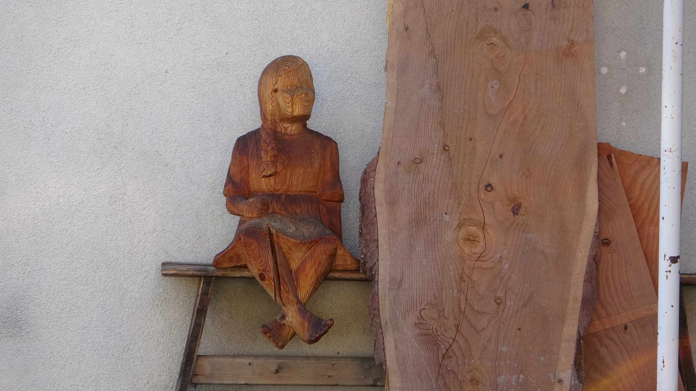 Geschnitzte sitzende Holzfigur auf einem kleinen Bänkchen neben einem unbearbeiteten Brett