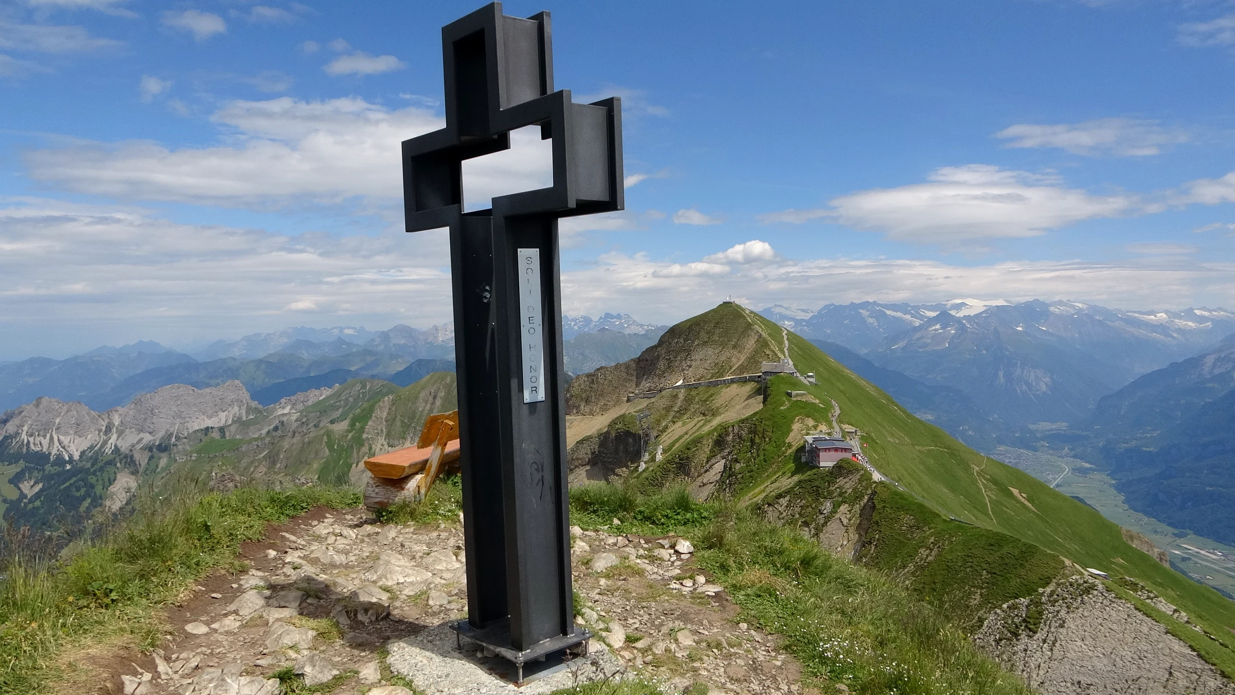 Schwarzes Gipfelkreuz aus Stahl auf dem Grat des Brienzer Rothorns, dahinter der Gipfel mit Bergstation