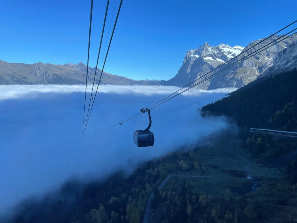 Eiger Express gondola above a sea of fog with the Wetterhorn rising behind under a blue sky