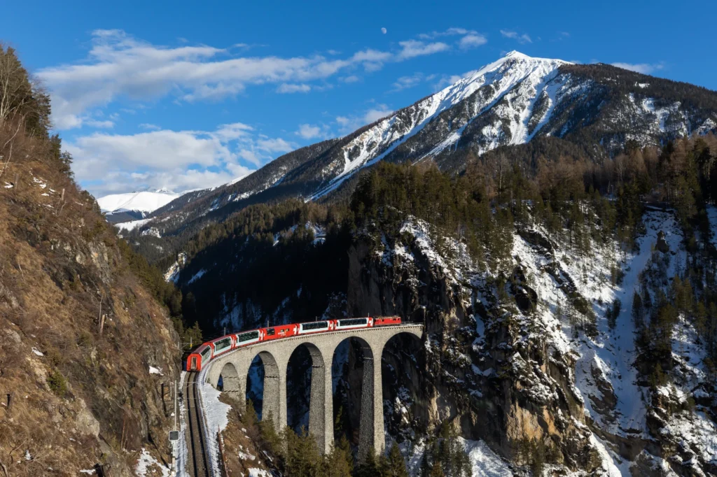 Glacier Express on the Landwasser Viaduct in winter with snow on the forest and a white peak behind