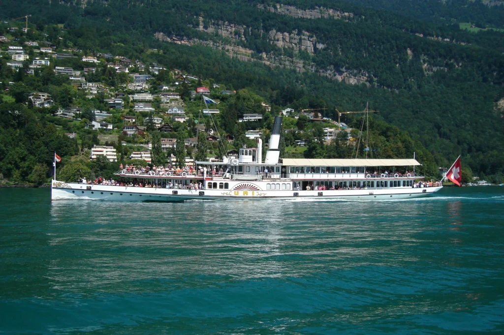 White paddle steamer Uri with passengers on deck on turquoise Lake Lucerne below a wooded hillside with villas