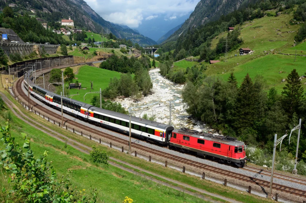 Red locomotive pulling the white Gotthard Panorama Express through a green valley next to a river