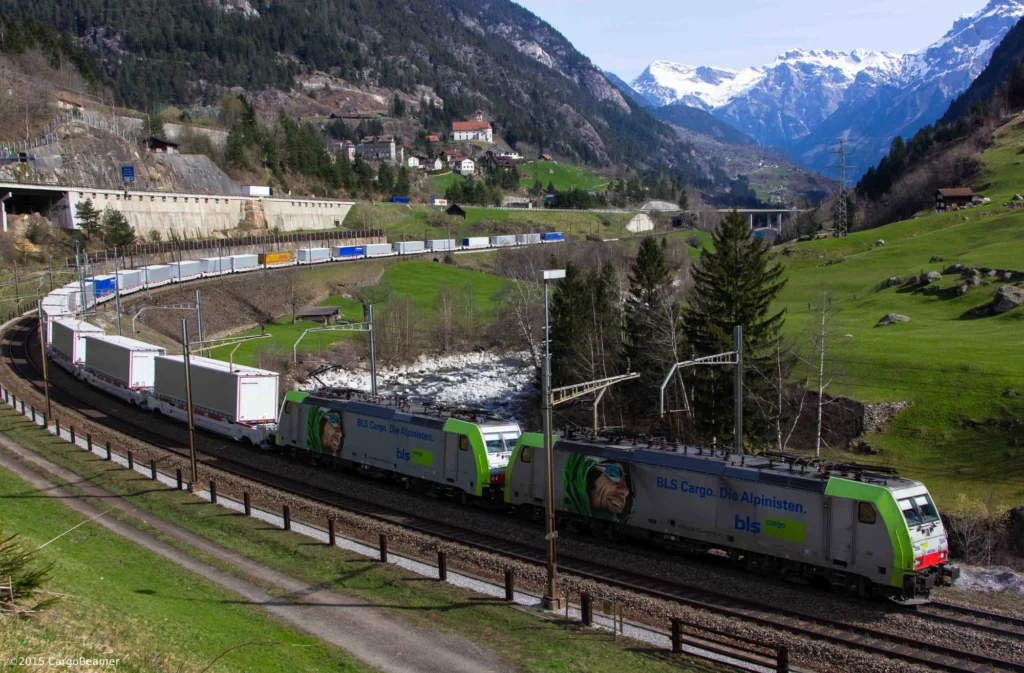 Train winding through the loops of the Gotthard line below the white church of Wassen with snowy peaks behind