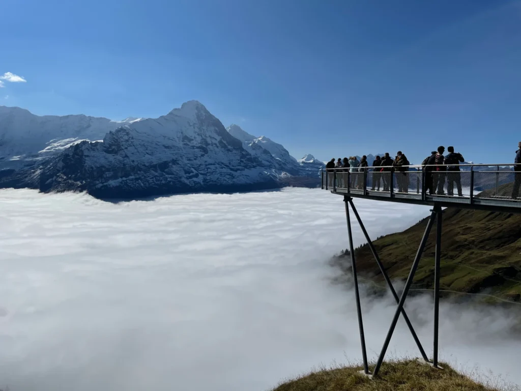 Plattform des First Cliff Walk über einem Nebelmeer, dahinter Eiger und verschneite Gipfel unter blauem Himmel