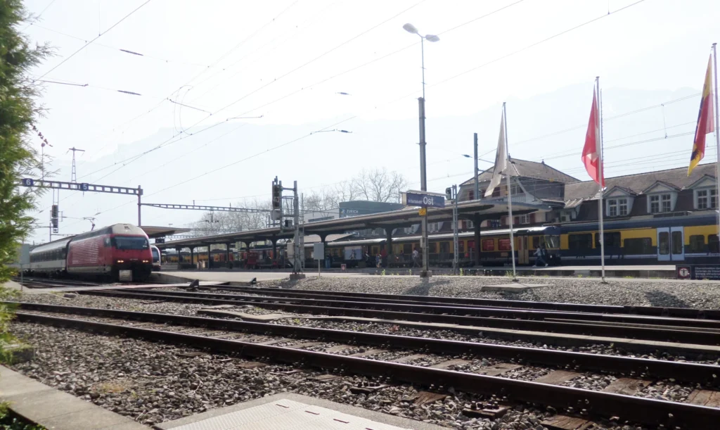 Interlaken Ost railway station from the east with tracks, a red SBB train and Swiss flags over the platforms