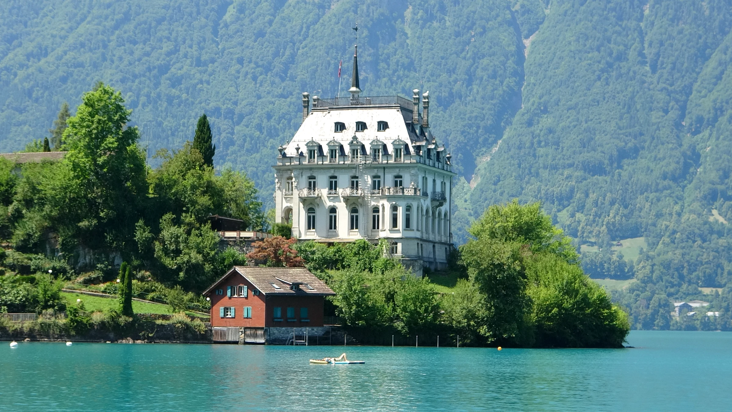 The white Seeburg castle on the wooded peninsula of Iseltwald with a red boathouse on Lake Brienz