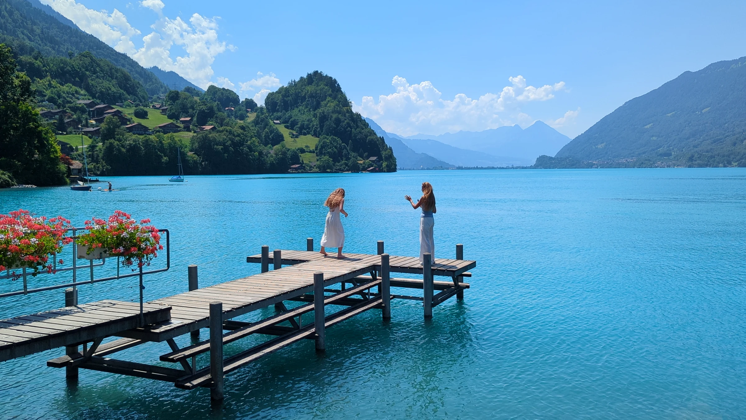Two women taking photos of each other at the end of the Iseltwald pier with the turquoise lake and mountains behind