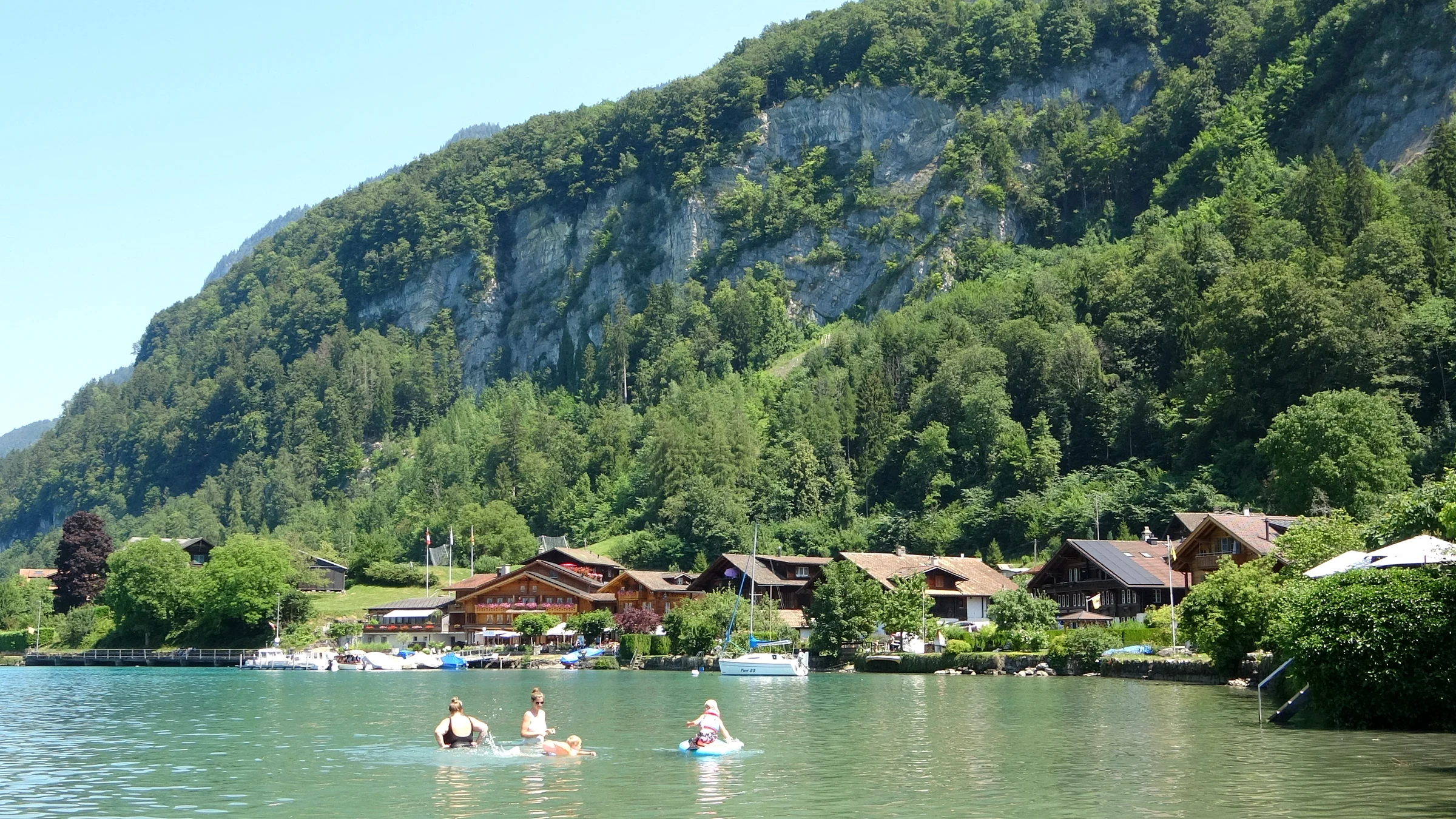 Swimmers and a paddleboarder in the turquoise bay of Iseltwald below a wooded cliff