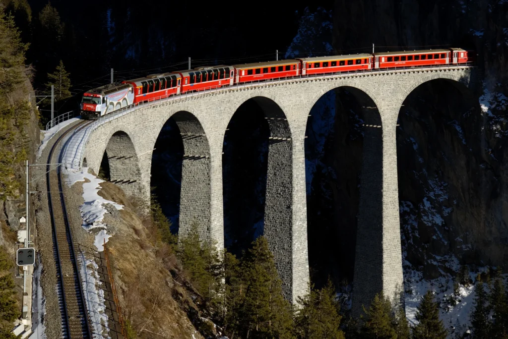 Roter Zug der Rhätischen Bahn auf den Steinbögen des Landwasserviadukts, Schnee auf den Felsen