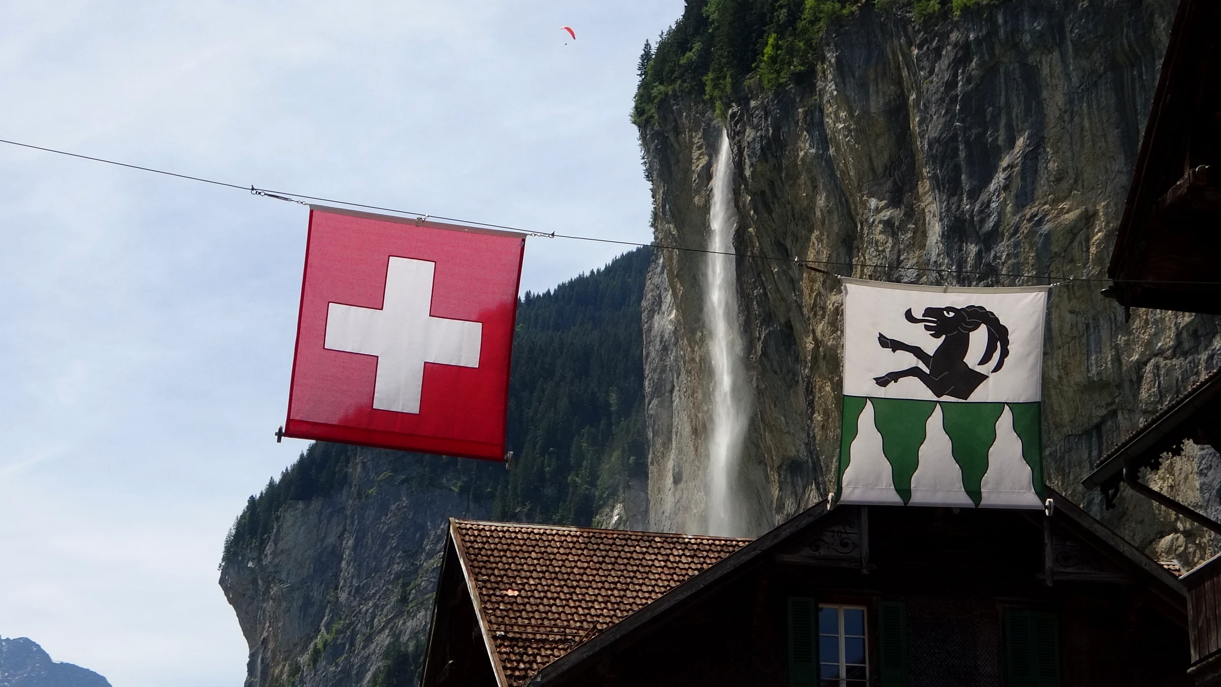 Staubbach Falls dropping down the cliff behind a chalet roof, with a Swiss flag and a local flag hanging over the street