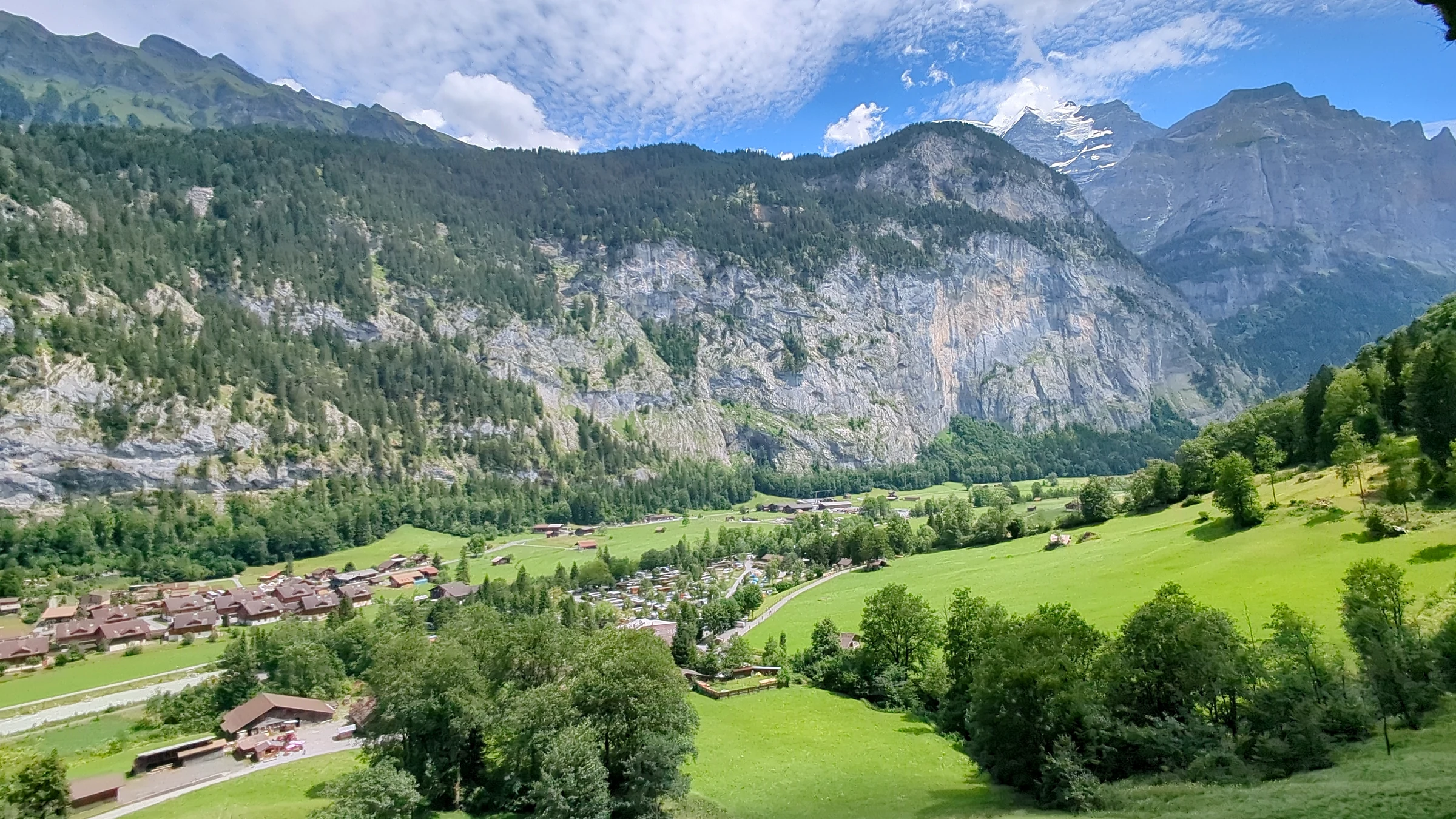 Lauterbrunnen Valley from the path above the village: green meadows, chalets and the long rock wall under a blue sky