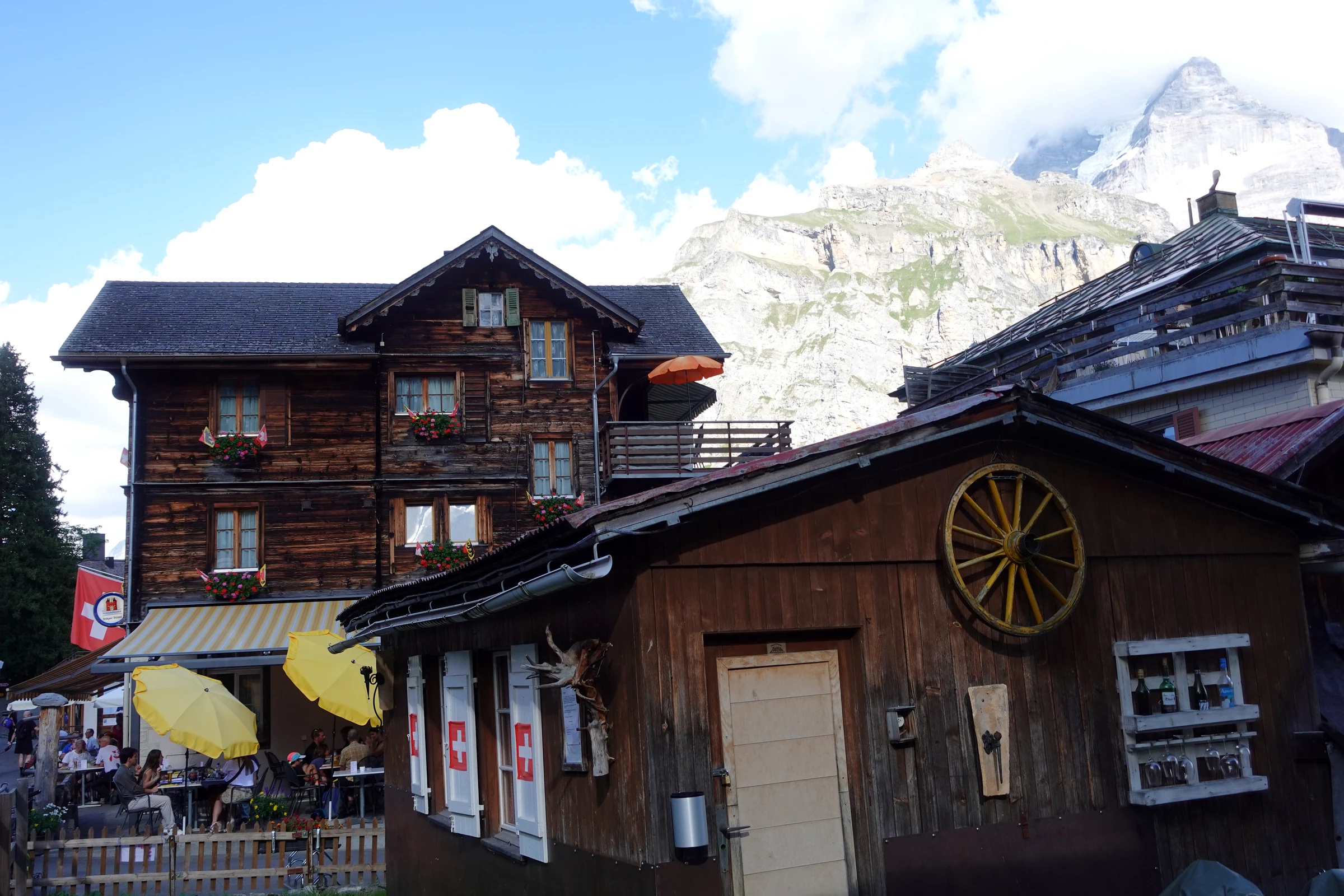 Old wooden chalets and a restaurant terrace with yellow umbrellas in car-free Mürren, rock peaks behind