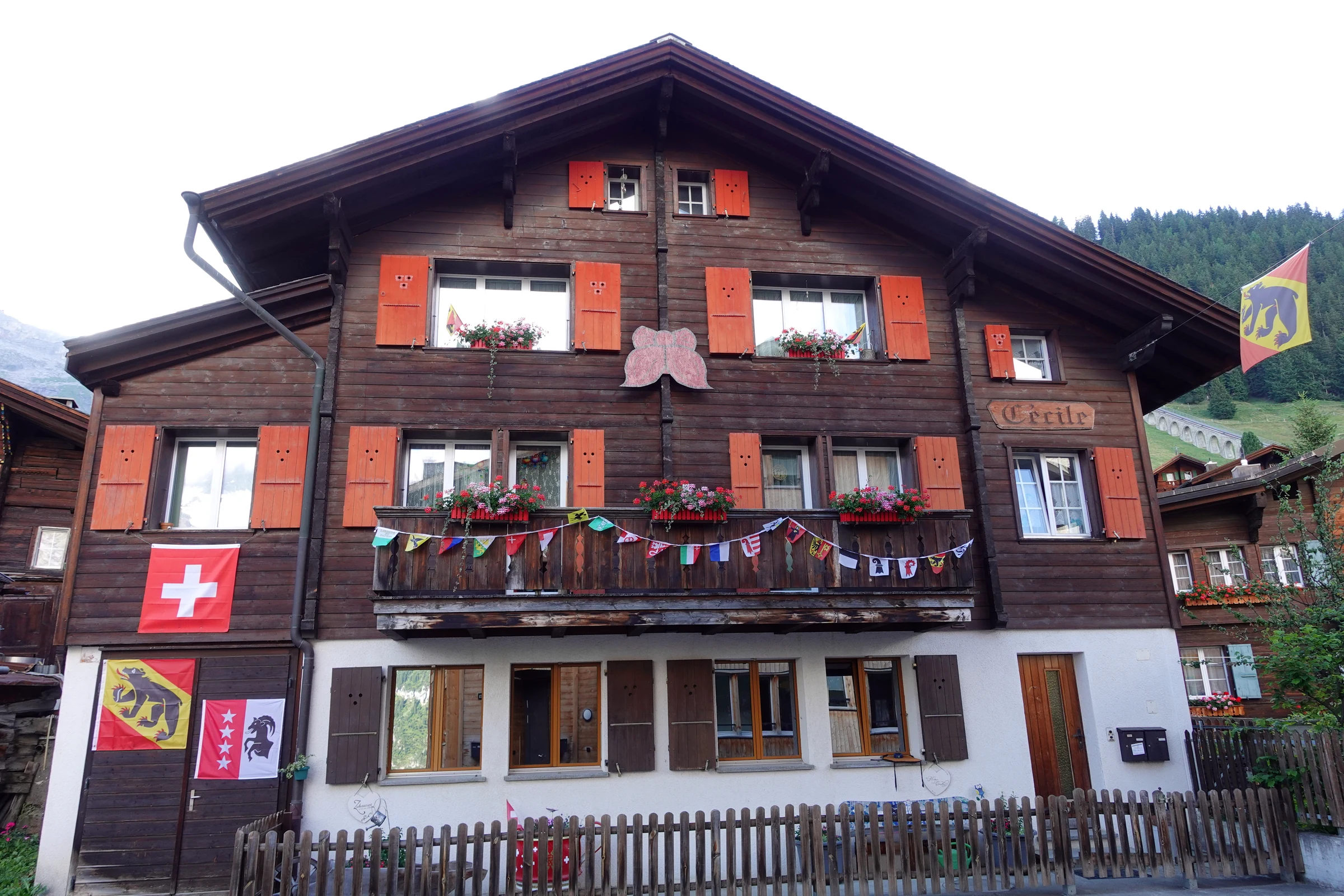 Chalet mit orangen Fensterläden, Schweizer Fahne, Berner Bär und Walliser Wappen, dahinter der Viadukt der Allmendhubelbahn