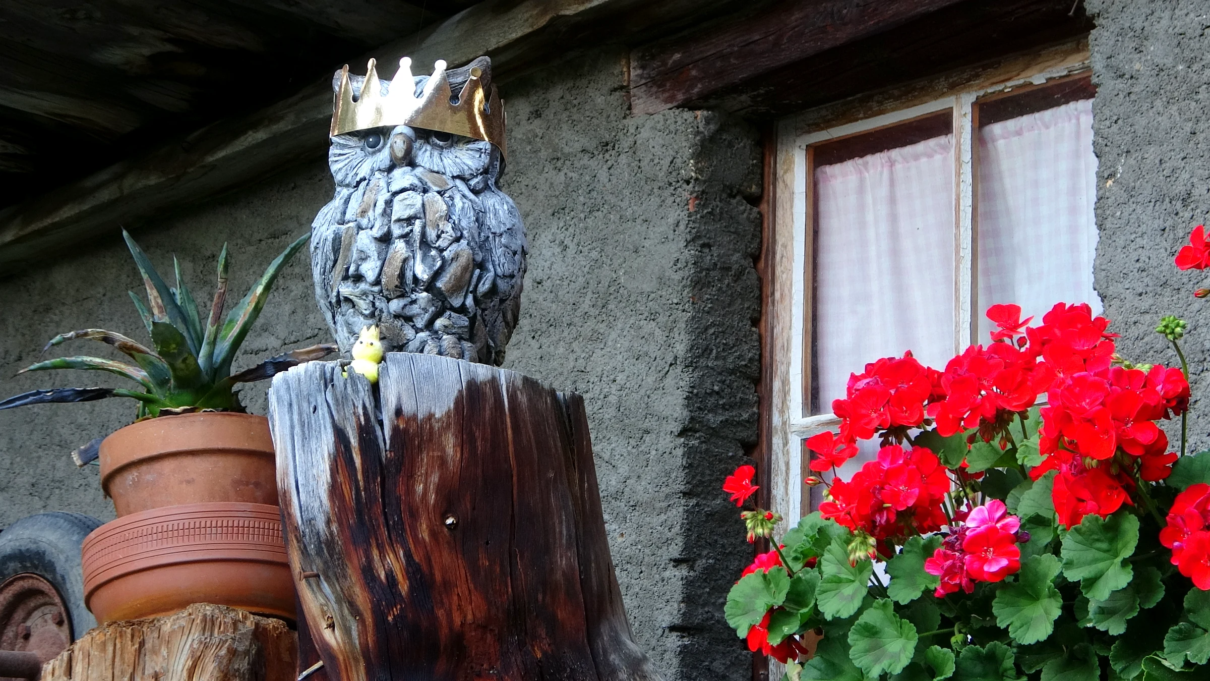 Geschnitzte Holzeule mit goldener Krone neben roten Geranien an einem Fenster in Mürren