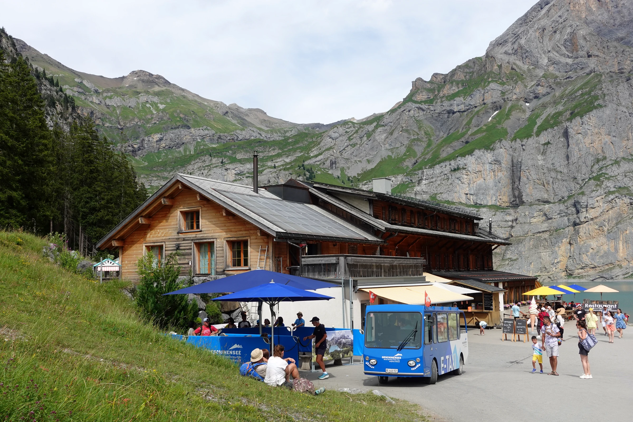Bergrestaurant Oeschinensee mit Sonnenschirmen auf der Terrasse und dem blauen Elektrotaxi davor