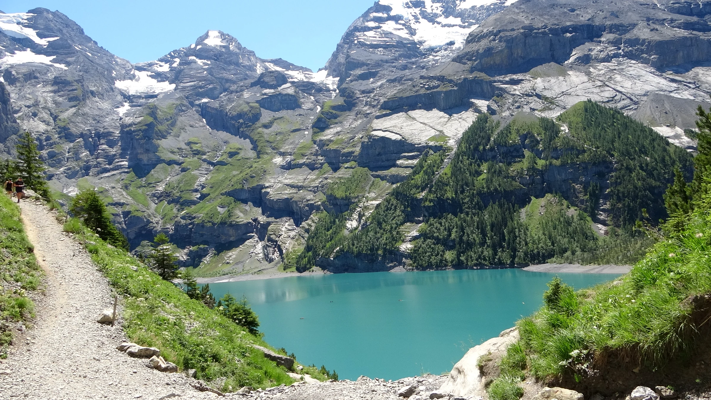 Schmaler Kiesweg an einem steilen Hang über dem Nordufer des Oeschinensees, dahinter vergletscherte Gipfel