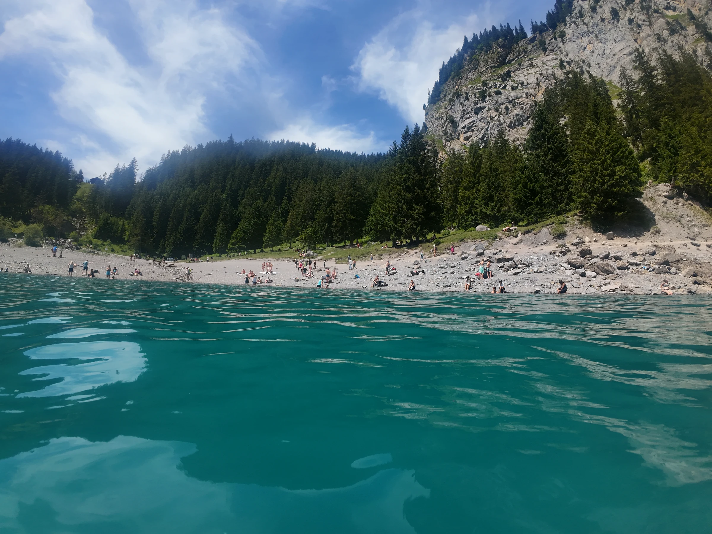 Türkises Wasser des Oeschinensees auf Höhe der Wasseroberfläche aus Schwimmersicht, dahinter ein voller Kiesstrand