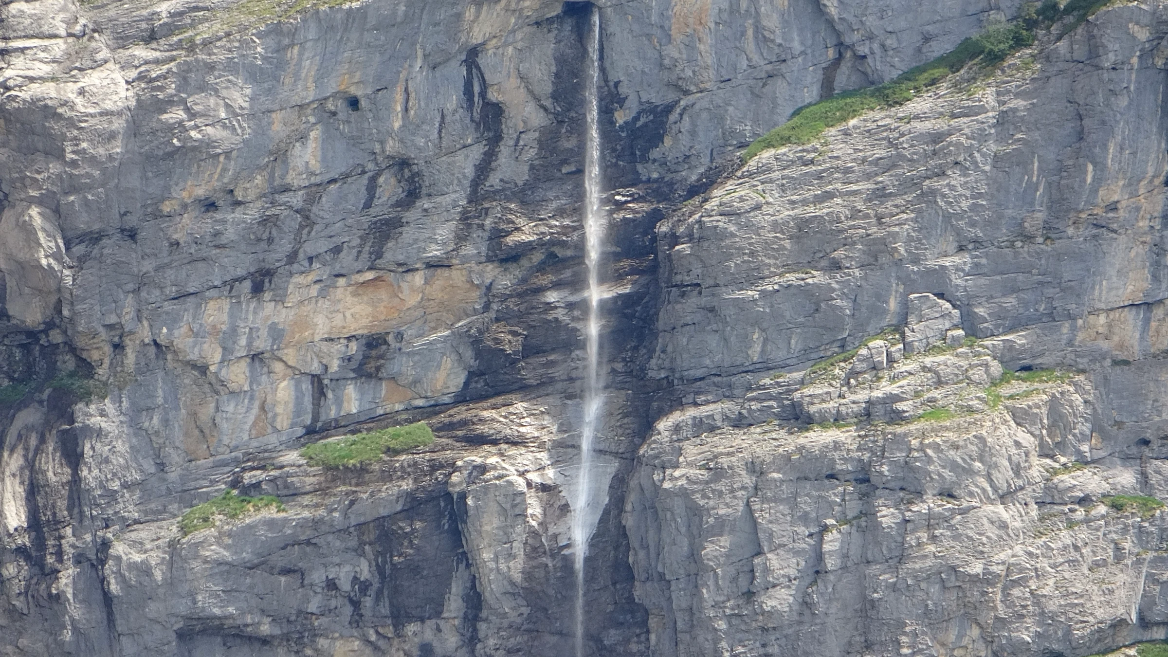 Schmaler Wasserfall stürzt über eine senkrechte graue Felswand in den Oeschinensee