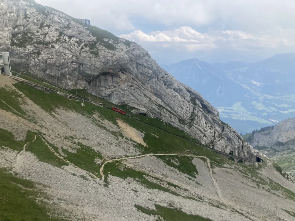 Red cogwheel train climbing a steep rocky slope below Pilatus Kulm with hiking paths in the scree