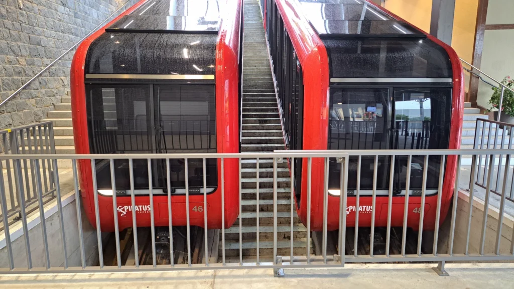 Zwei rote Triebwagen der Pilatusbahn mit den Nummern 46 und 48 nebeneinander in einer Station, dazwischen eine Treppe