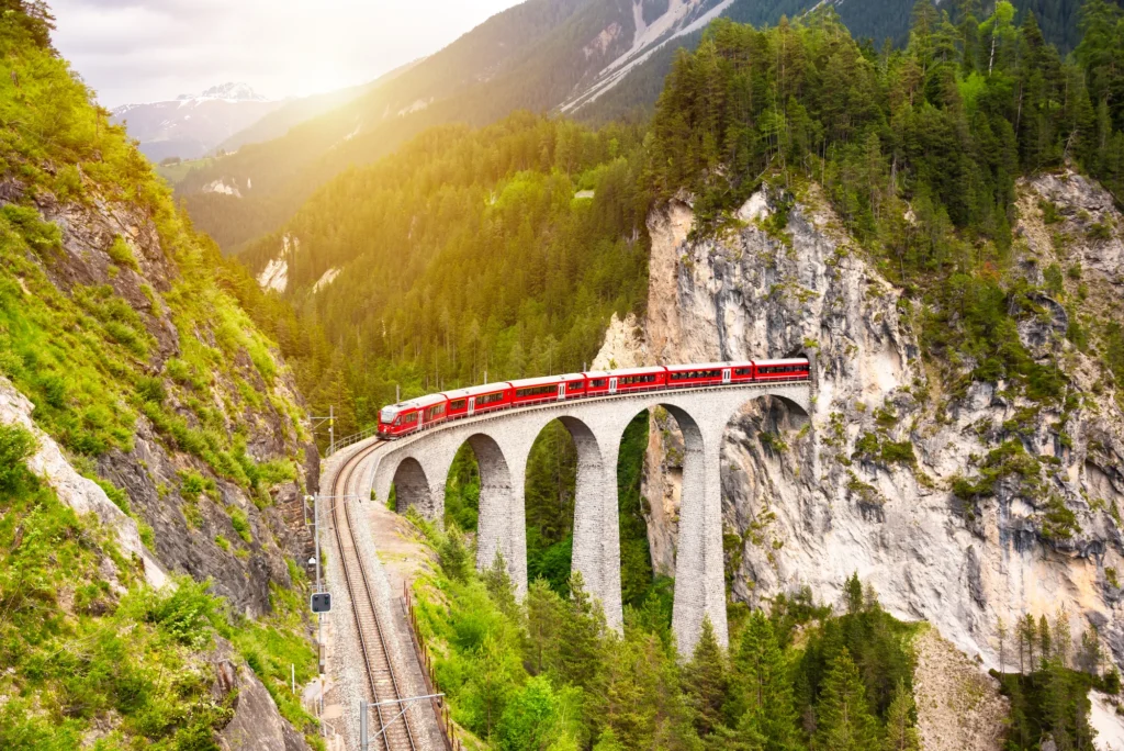 Red train crossing a stone viaduct in a green mountain valley in evening light