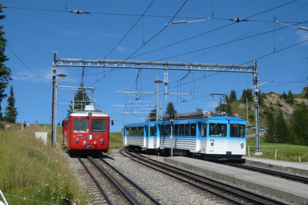 A red and a blue Rigi cogwheel train side by side on parallel tracks below Rigi Kulm under a blue sky