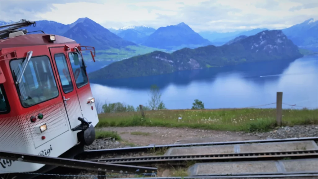 Front of a red and white Rigi train at a station high above Lake Lucerne with the Bürgenstock and mountains behind