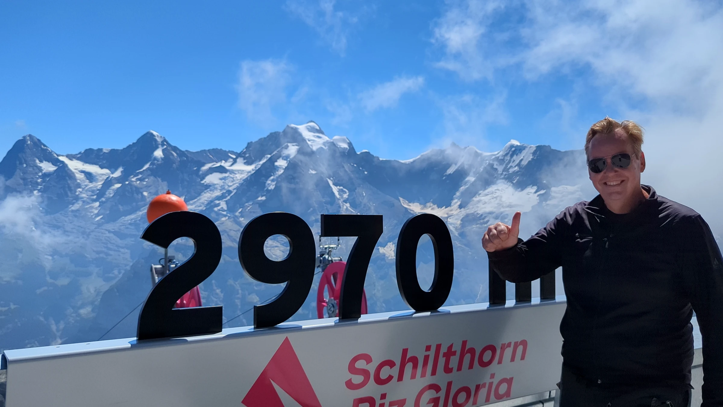 Autor Alexander Haselhoff mit Daumen hoch neben dem 2970-m-Schild auf dem Schilthorn-Gipfel, dahinter Eiger, Mönch und Jungfrau