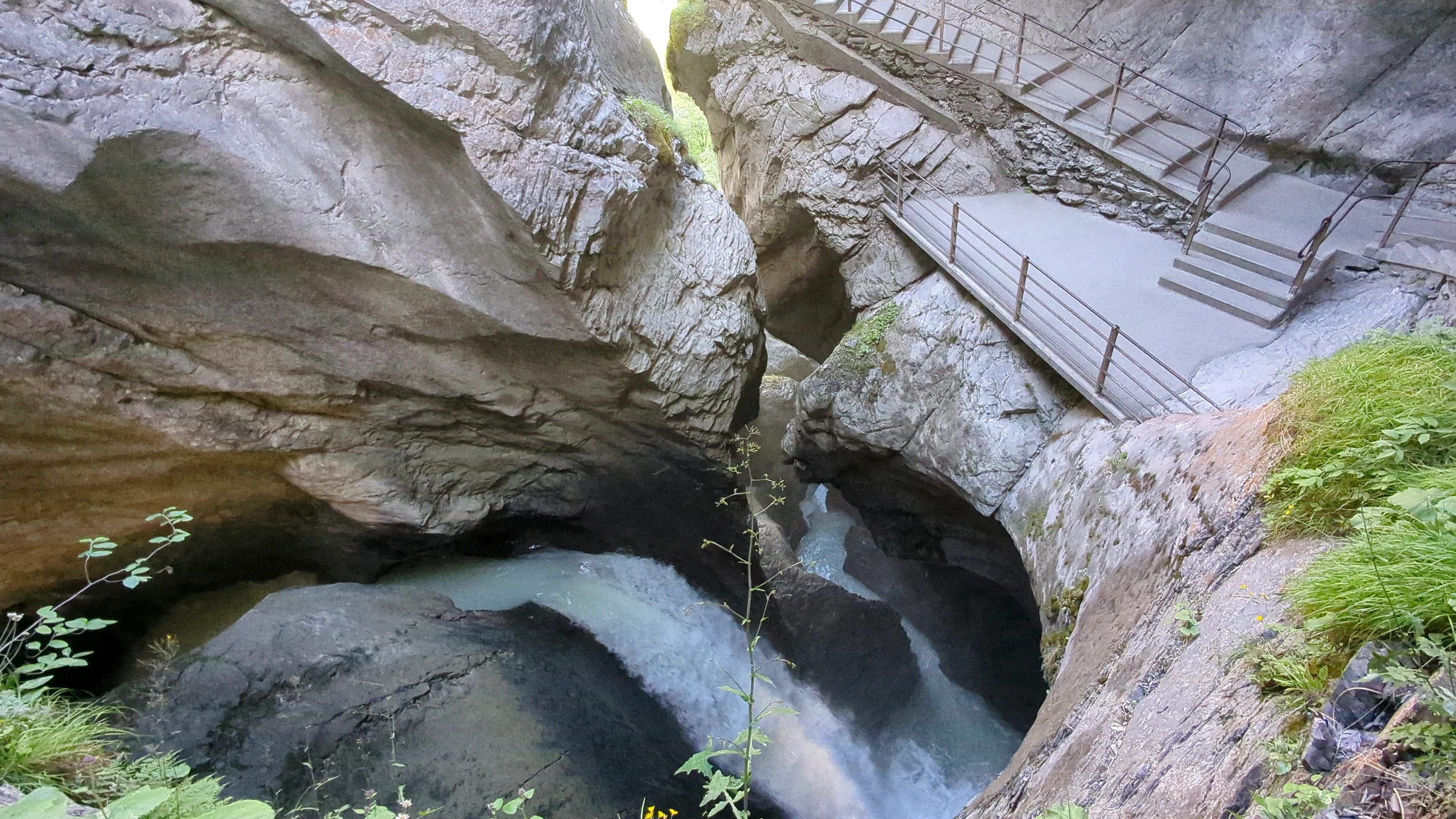 Gletscherwasser schraubt sich durch ein rundes Felsbecken der Trümmelbachfälle, darüber Treppe und Steg