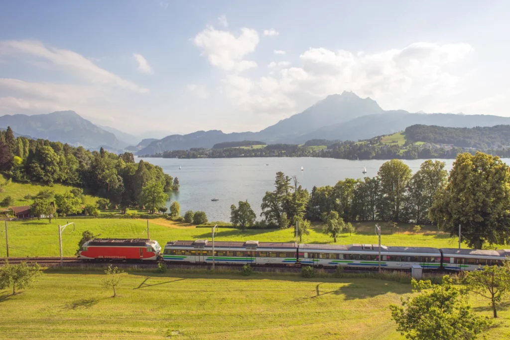 Voralpen-Express train passing green meadows on the shore of Lake Lucerne with mountains behind